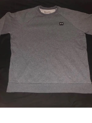 Grå sweatshirt från Under Armour - Snygg grå sweatshirt från Under Armour med rund halsringning och diskret logga på bröstet. Tröjan har långa ärmar och ribbade muddar vid ärmslut och nederkant. Perfekt för en avslappnad och sportig stil.