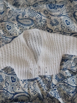 Vit stickad kofta med knappar till en bebis - Söt vit stickad kofta med v-ringning och dekorativa hålmönster. Koftan har långa ärmar och stängs med små vita knappar framtill. Perfekt för lager på lager och ger en mysig vibe till din outfit.