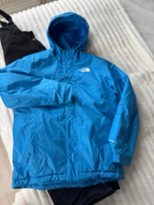 Skidställ med blå jacka och svarta byxor  från The North Face - Blå skidjacka från The North Face med huva och dragkedja. Jackan har en stilren design med logga på bröstet och är tillverkad i ett slitstarkt material som passar perfekt för utomhusaktiviteter. Tillhörande termobyxor i svart med logga på benet. Klassisk passform och praktisk för skidåkning eller vinteräventyr. Passar ungdom 150-160 lång.
