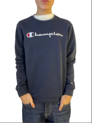 Mörkblå Champion sweatshirt - Mörkblå sweatshirt från Champion med broderad logga i vitt, rött och blått på bröstet. Tröjan har rund halsringning, långa ärmar och ribbade muddar vid ärmslut och nederkant. Perfekt för en avslappnad och sportig stil.