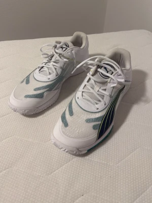 Vita Puma sneakers med blå detaljer - Snygga vita sneakers från Puma med blå och gröna detaljer på sidorna och meshmaterial för extra andningsförmåga. Skorna har klassisk snörning och en sportig design som passar perfekt till en aktiv livsstil.