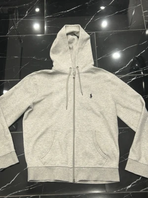 Ralph Lauren zip hoodie - Säljer en stilren och bekväm zip-hoodie från Polo Ralph Lauren i ljusgrå färg. Klassisk modell med dragkedja, huva och broderad Polo-logga på bröstet. Tillverkad i mjukt och skönt material av hög kvalitet. Tröjan är nästan oanvänd och i mycket fint skick. Det finns dock en liten fläck på insidan, som beroende på ljus kan svagt även ses på utsidan (se bild). Men det är inget  som påverkar funktion eller passform. Det finns även en QR kod i tröjan som går att skanna för att se att den är äkta