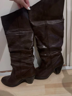 Bruna höga läderstövlar med klack - Säljer ett par mörkbruna stövlar i äkta läder med hög, vikbar skaft och markerad klack. Stövlarna har en slouchy passform och en klassisk spetsig tå. Perfekta för dig som vill ha en tidlös och cool look. De är använda men fint skick se bilder.Går att vika ned Ingen dragkedja  Storlek 37 Fint men använd skick (se bilder) Märket är Colore per favore ,Lite fodrade