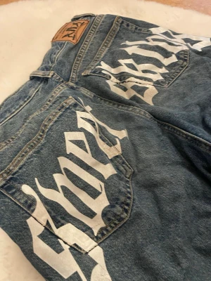 Blåa baggy jeans med tryck på baksidan - Säljer ett par blåa jeans från SKTBS med stort vitt tryck i gotisk stil över bakfickorna. Jeanssn är baggy. Texten på baksidan har spruckit lite vid fickan men det märks inte. Perfekta för en streetwear-look. Köpte jeansen för 500kr!