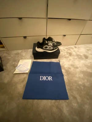 Svarta Dior B30 sneakers - Snygga svarta Dior B30 sneakers med vita och grå detaljer samt tydlig Dior-logga på sidan. Skorna har en chunky vit sula och vita snören. Tillverkade i en mix av mesh och läder för en modern look. Perfekta för dig som vill sticka ut med exklusiva sneakers.