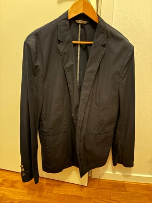 Marinblå kavaj från Hugo Boss - Snygg marinblå kavaj från Hugo Boss i italienskt tyg. Klassisk modell med två framfickor, knappar vid ärmslut och diskret innerfoder med mönster. Perfekt för en stilren look.