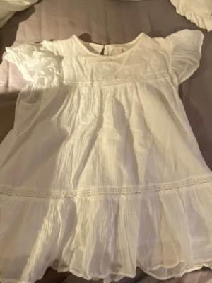 Vit luftig blus med volanger - Supersöt vit blus i tunn, luftig bomull med korta ärmar och volangdetaljer. Blusen har rund halsringning och dekorativa spetsband. Perfekt för en fräsch och somrig look.