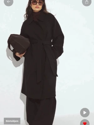 Svart kappa med knytskärp - Helt ny, aldrig använd. Stilren svart kappa med klassisk krage och knytskärp i midjan. Kappan har raka ärmar och en rak passform som ger en clean look. Perfekt för dig som gillar minimalistisk stil. Materialet ser ut att vara ullblandning.