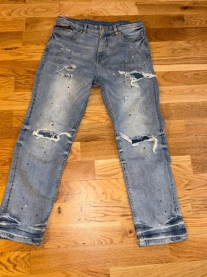 Ljusa slitna jeans Supply & Demand - Säljer ett par ljusblå jeans från Supply & Demand med slitningar och hål på benen samt färgstänk för en trendig look. Klassisk femficksmodell och raka ben. Perfekta för dig som gillar streetstyle och vill sticka ut lite extra.