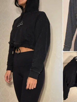 Svart cropped hoodie Couture Club - Säljer en svart cropped hoodie från The Couture Club med huva och dragsko i midjan. Hoodien har en stor magficka och vit broderad logga på bröstet. Tillverkad i mjukt sweatshirtmaterial, perfekt för en chill och trendig look.