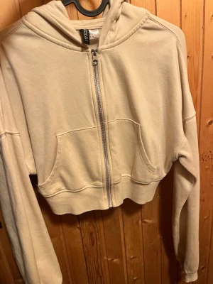 Beige cropped hoodie med dragkedja - Snygg beige cropped hoodie från H&M med dragkedja framtill och två fickor. Hoodien har huva och ribbade muddar vid ärmslut och nederkant. Perfekt för en avslappnad och trendig stil.