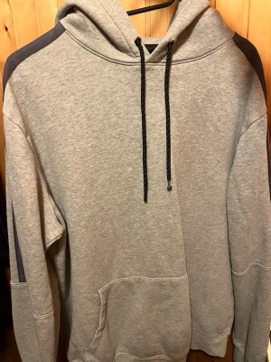 Grå hoodie med svarta detaljer - Snygg grå hoodie med svarta dragsnören och mörkare grå paneler längs ärmarna. Tryck med koordinater på ärmarna ger en unik touch. Stor känguruficka framtill och mjukt material, perfekt för chilla dagar.