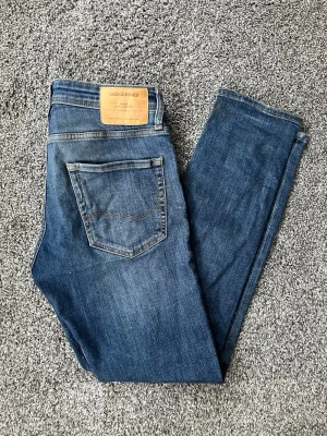 Blå slim fit jeans från Jack & Jones - Snygga blå slim fit jeans från Jack & Jones med klassisk femficksdesign och läderpatch bak i midjan. Jeansen har en lätt tvättad look och smal passform som sitter tajt längs benen. Perfekta för en avslappnad och trendig stil.