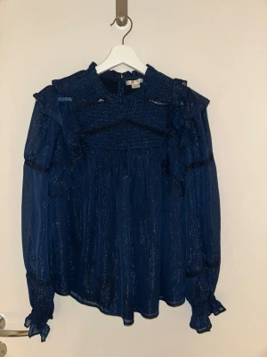 Mörkblå volangblus med glitter - Supersnygg mörkblå blus med volanger och subtilt glittrande tyg. Blusen har långa puffärmar med resår och volangdetaljer vid axlar och ärmslut. Materialet är lätt och transparent, perfekt för att sticka ut. Hög hals och avslappnad passform.