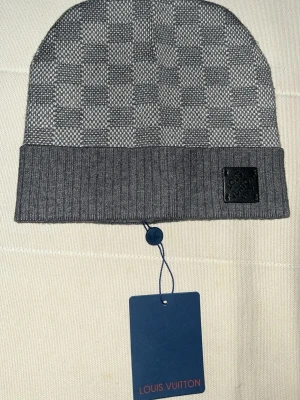 Grå Louis Vuitton mössa - Snygg grå mössa Louis Vuitton med klassiskt rutmönster och ribbad uppvikt kant. Märkets logga sitter på en svart patch framtill. Perfekt accessoar för att lyfta din stil under kyliga dagar.