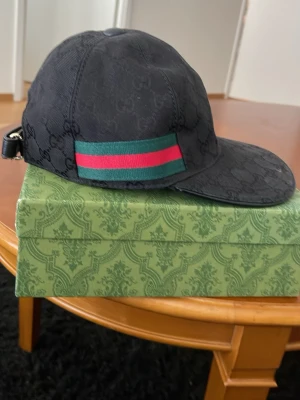 Svart Gucci keps med grön-röd rand - Säljer en svart keps från Gucci med diskret GG-mönster i tyget och klassiska grön-röda ränder på sidan och baktill. Justerbar rem bak i läder och böjd skärm. Perfekt för dig som vill ha en stilren och lyxig accessoar.
