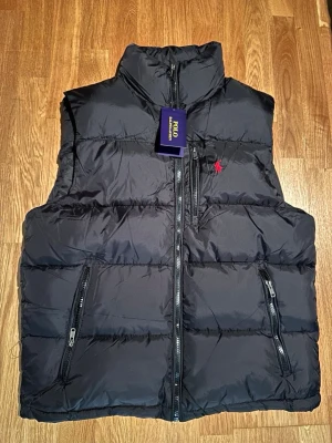 Svart dunväst från Polo Ralph Lauren - Snygg svart dunväst från Polo Ralph Lauren med hög krage, tre dragkedjeförsedda fickor och klassisk röd logga på bröstet. Västen har en puffig design och är tillverkad i ett glansigt material som ger en modern look. För mer bilder skriv gärna. Pris går att diskutera.