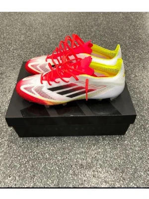 Adidas  fotbollsskor PRO  - Snygga fotbollsskor från Adidas med vit bas, röda detaljer och gula inslag. Skorna har svarta ränder på sidan och röda snören. Designade för bra grepp på planen och tillverkade i syntetmaterial för lätt känsla och snabbhet.