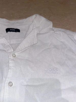 Vit kortärmad skjorta från Hugo Boss - Fräsch vit skjorta från Hugo Boss med korta ärmar och klassisk krage. Skjortan har knäppning framtill och diskret broderad logga på bröstet. Perfekt för en stilren och clean look.