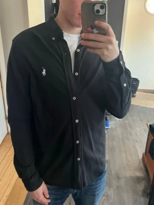 Svart skjorta från Ralph Lauren - Klassisk svart skjorta i riktigt fett featherweight mesh material från Ralph Lauren med vit broderad logga på bröstet. Skjortan har långa ärmar, vita knappar och normal passform. Perfekt till jeans eller chinos för en clean och stilren look.