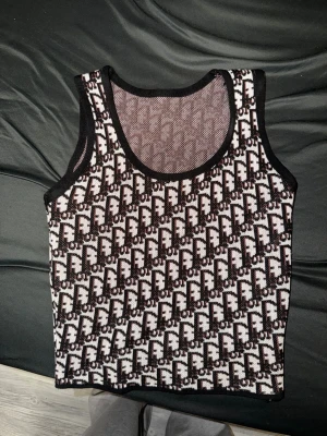 Svartvit mönstrad blus från Fendi - Säljer en snygg svartvit blus från Fendi med ett grafiskt F-mönster över hela plagget. Blusen är ärmlös och har svarta kanter runt hals och ärmhål. Materialet är stickat och känns mjukt mot huden. Perfekt för dig som gillar designerplagg med tydlig logga.