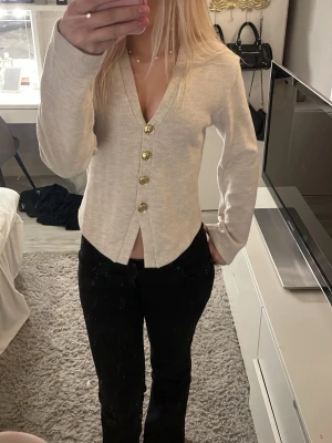 Beige kofta med guldknappar - Snygg beige kofta med djup V-ringning och fyra dekorativa guldknappar framtill. Koftan har lång ärm och är tillverkad i ett mjukt, ribbat material som ger en stilren look. Perfekt att styla med jeans eller kjol för en trendig outfit. 