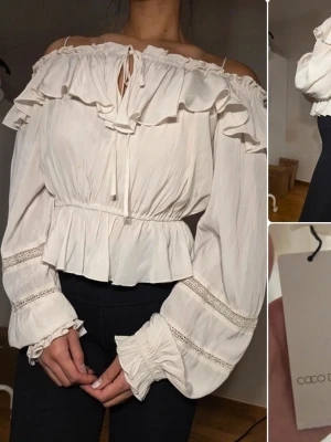 Off shoulder blus med volanger Coco Deal - Supersöt off shoulder blus från Coco Deal i krämvitt. Blusen har volanger vid halsringningen, långa puffärmar med spetsdetaljer och resår vid ärmslut och midja. Tillverkad i 100% bomull, luftig och perfekt för en trendig look.