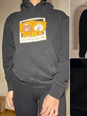 Svart hoodie LINE FRIENDS med tryck - Svart hoodie från LINE FRIENDS x Uniqlo med gulligt tryck av två figurer i djurdräkter och texten 'Let's celebrate together!'. Klassisk känguruficka och huva. Mjuk bomullskänsla och relaxed fit. Perfekt för dig som gillar söta motiv och streetwear.