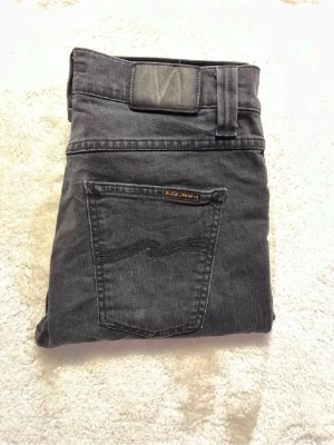 Svarta Nudie Jeans - Säljer ett par svarta jeans från Nudie Jeans med klassisk femficksdesign och raka ben. Jeansen har en diskret logga på bakfickan och är tillverkade i mjukt denimtyg som sitter snyggt. W29 L32
