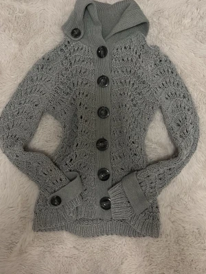 Grå stickad kofta från STOCKH LM - Snygg grå stickad kofta från STOCKH LM med stora svarta knappar framtill. Koftan har ett unikt hålmönster, ribbade muddar och hög krage. Perfekt för dig som gillar en trendig och mysig stil.