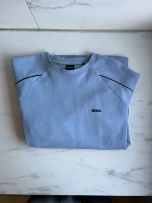 BOSS tröja  - Ljusblå sweatshirt från BOSS med svarta detaljer längs ärmarna och diskret BOSS-logga på bröstet. Storlek L, fint skick !