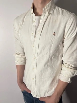 Vit skjorta från Ralph Lauren - Klassisk vit skjorta från Ralph Lauren med broderad logga på bröstet. Skjortan har lång ärm, knappar framtill och en snygg krage. Tillverkad i bomull för en skön och stilren känsla. Perfekt till jeans eller chinos.