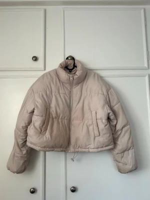 Beige cropped pufferjacka - Säljer en beige cropped pufferjacka med hög krage och dragkedja framtill. Jackan har två sidofickor och justerbar dragsko i nederkant. Perfekt för kalla vinterdagar och ger en trendig siluett. Materialet är quiltat och ger en fluffig känsla.