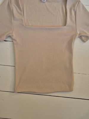 Beige ribbad topp med fyrkantig ringning - Trendig beige topp med ribbad struktur och fyrkantig ringning. Kortärmad och croppad modell som passar perfekt till både jeans och kjol. Enkel men stilren design som är lätt att matcha.