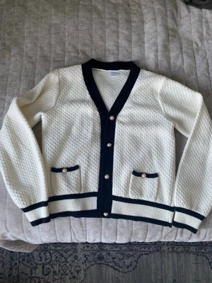 Vit kofta med svarta detaljer Lindex - Stilren vit kofta från Lindex med svarta ribbade kanter och knappar med pärldetalj. Koftan har två små fickor framtill och ett diskret rutmönster i stickningen. Perfekt för dig som gillar klassisk preppy stil.