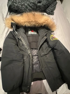 Svart parkas från Zavetti Canada - Snygg svart parkas från Zavetti Canada med stor huva och fuskpäls. Jackan har ett broderat märke på ärmen och dragkedja framtill. Perfekt för kalla dagar och har en klassisk, stilren look. Priset kan diskuteras!
