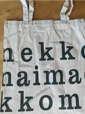 Marimekko tygkasse / shoppingpåse / tygväska - Stilren shoppingväska i beige bomullstyg med stora svarta bokstäver tryckta på framsidan. Väskan har två långa handtag och är perfekt för att bära böcker, mat eller annat du behöver. Enkel och trendig design som passar till det mesta.