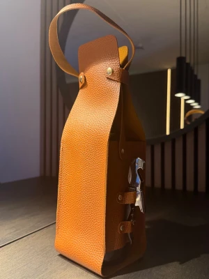 Läderfodral för vin- & champagneflaska - Elegant flaskbärare i exklusivt läder, framtagen för att bära och presentera vin- eller champagneflaskor med stil. En premium accessoar som passar utmärkt som present, vid middagar, utflykter eller som en dekorativ detalj i hemmet.  Passar standardstorlek på vin- och champagneflaskor.  ⚠️ OBS: Flaska & korkskruv medföljer inte.