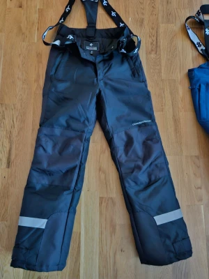 Svarta täckbyxor Cross Sportswear - Svarta täckbyxor från Cross Sportswear med justerbara hängslen och reflexdetaljer på benen. Byxorna har två fickor fram och förstärkta knän. Perfekta för vinterns alla äventyr i snön.  OBS! Fraktfritt i 7 dagar från annonsen startats enligt Plick!