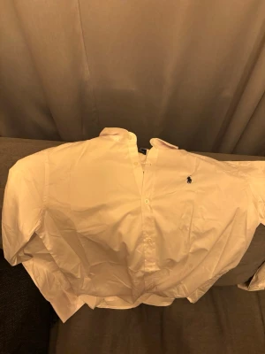 Vit skjorta från Ralph Lauren - Klassisk vit skjorta från Ralph Lauren med broderad logga på bröstet. Skjortan har lång ärm, knappar framtill och en tidlös design som passar till många olika stilar. Tillverkad i bomull för en bekväm känsla.