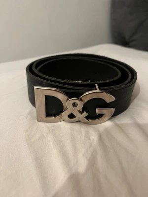 Svart skärp från Dolce & Gabbana - Stilrent svart skärp från Dolce & Gabbana med en stor, silverfärgad D&G-logga som spänne. Skärpet är gjort i skinn och passar perfekt för att lyfta vilken outfit som helst med en lyxig touch. 