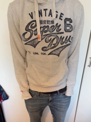 Beige hoodie från Superdry - Snygg beige hoodie från Superdry med stor svart/mörkblå logga och tryck på bröstet. Klassisk känguruficka framtill, dragsko i huvan och ribbade muddar. Perfekt för en avslappnad streatwear look.