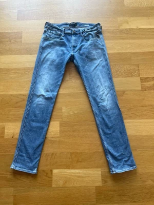 Blåa raka replay anbass jeans med slitningar W34 L34 L34 - Säljer ett par klassiska blå jeans med raka ben och lätt slitna detaljer framtill. Jeansen har fem fickor, bälteshällor och knappgylf. Materialet är denim i bomull och passformen är avslappnad men ändå snyggt åtsittande.