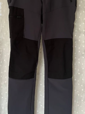 Svarta och mörkgrå friluftsbyxor - Snygga friluftsbyxor i svart och mörkgrått med förstärkta partier över knän och bak. Byxorna har dragkedjeförsedda fickor och är tillverkade i slitstarkt syntetmaterial, perfekta för utomhusaktiviteter och äventyr. Stl 158.