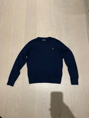 Mörkblå tröja från Ralph Lauren - Mörkblå långärmad tröja från Ralph Lauren med klassisk rund halsringning och den ikoniska broderade loggan i orange på bröstet. Tröjan har en stilren och enkel design, perfekt för en clean look.