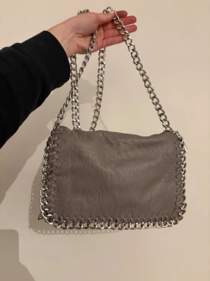 Grå axelväska med kedjedetaljer - Säljber nu min snygga gråa axelväska i mjukt, strukturerat konstläder med silverfärgad kedja som axelrem och runt kanterna. Den är i bra skick. 