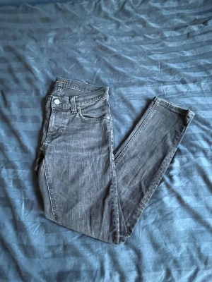 Svarta skinny jeans från Nudie Jeans - Säljer ett par svarta jeans från Nudie Jeans med klassisk femficksdesign och diskret broderad bakficka. Jeansen har en mörk tvätt och är tillverkade i stretchig bomull för en skön passform. Perfekta för dig som gillar en stilren och enkel look.