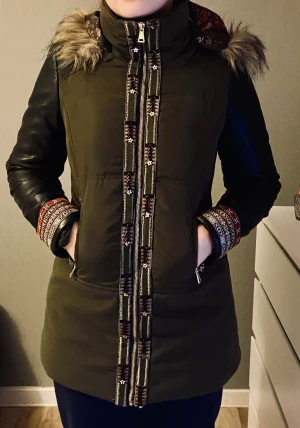 Grön parkajacka från Desigual med päls - Snygg grön parkajacka från Desigual med svarta skinnärmar, fuskpäls på huvan och broderade detaljer vid dragkedjan och ärmsluten. Fodret har blommigt mönster och jackan har praktiska fickor med dragkedja. Perfekt för dig som gillar unika detaljer. Otroligt varm o härlig jacka. 