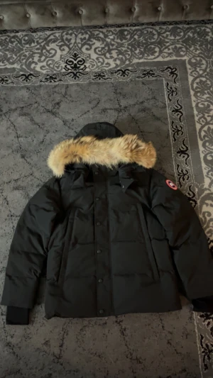Svart dunjacka Canada Goose med päls - Svart dunjacka från Canada Goose med ikonisk röd och vit logotyp på ärmen. Jackan har huva med beige päls, långa ärmar och knäppning med både dragkedja och knappar framtill. Perfekt för kalla vinterdagar och har en klassisk, stilren look.