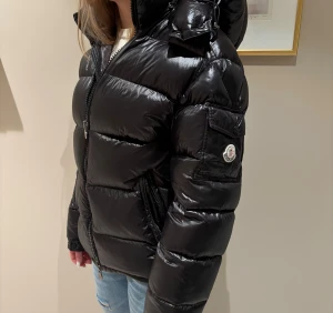 Svart Moncler* Maya pufferjacka stl 1 XS/S - Säljer en mycket fin och varm svart pufferjacka från Moncler med glansig finish och huva. Köpt på Vinted. *Jag kan inte garantera äkthet då jag saknar originalkvitto, men jag har gjort de rekommenderade kontrollerna utan att hitta något misstänkt. Säljs därför till ett lägre pris. Det finns en skada fram på jackan som är lagad av skräddare, mkt snyggt. Se bild. Storlek 1 som jag bedömer som S/XS